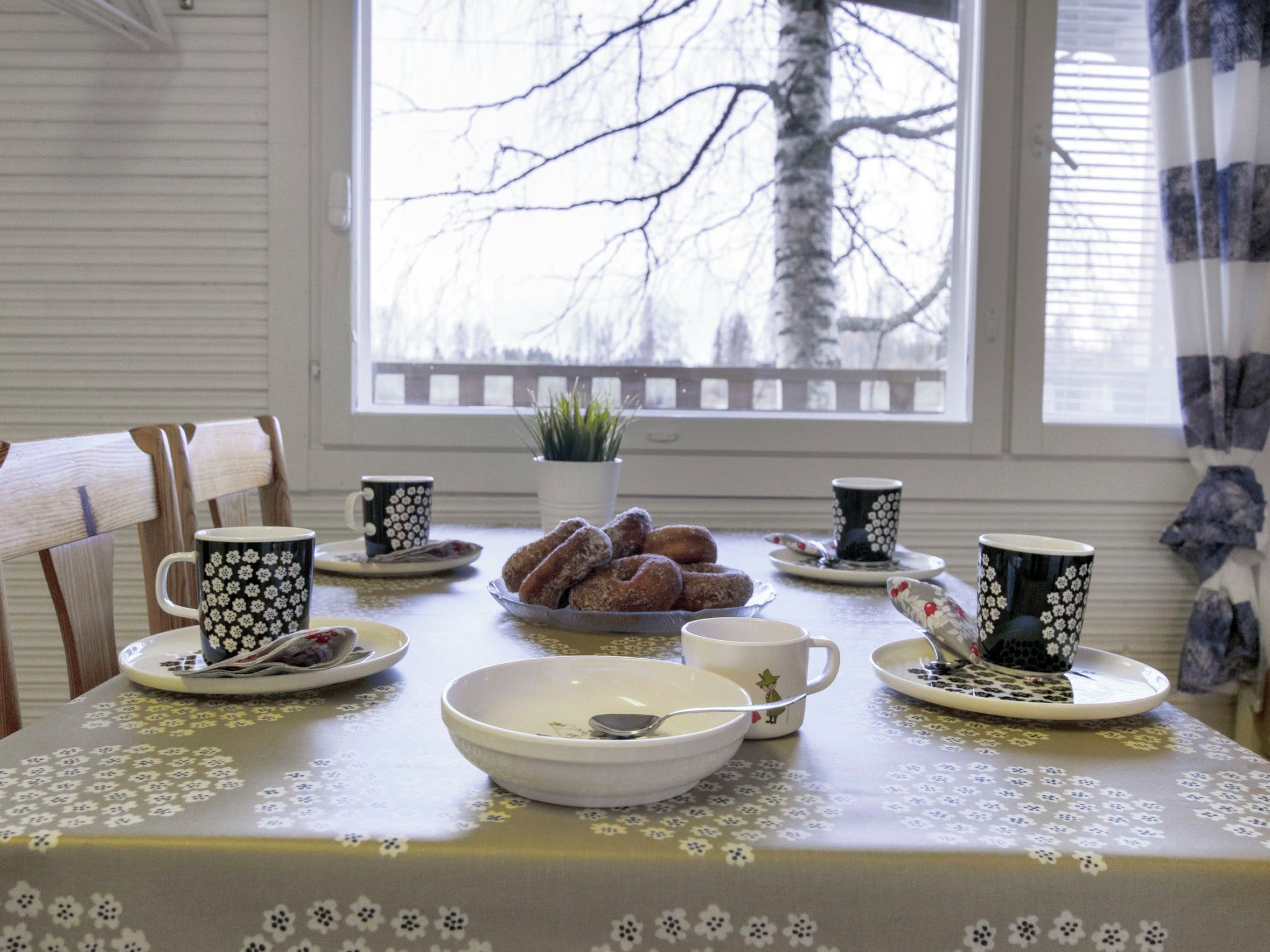 A dining table with floral tablecloth, black and white patterned cups, a bowl, and a plate of pastries.