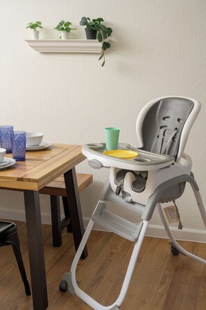 Een eetruimte met een houten tafel, een kinderstoel met dienblad, en een plank met potplanten.