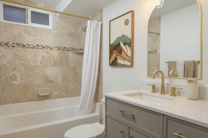 A bathroom with a bathtub, toilet, sink, and a mirror with a painting.