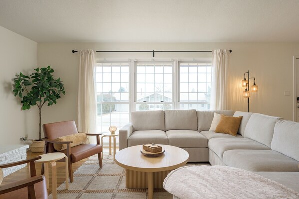 A modern living room with a beige sofa, a wooden coffee table, a potted plant, and a view of the outside through large windows.