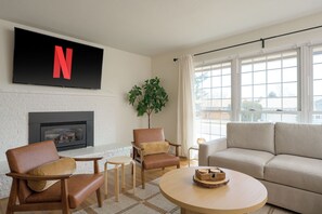 Een woonkamer met een open haard, een aan de muur bevestigde televisie, een bank, twee fauteuils en een salontafel.