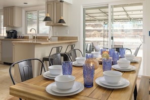A dining area with a wooden table set for four, surrounded by chairs, with a kitchen in the background.
