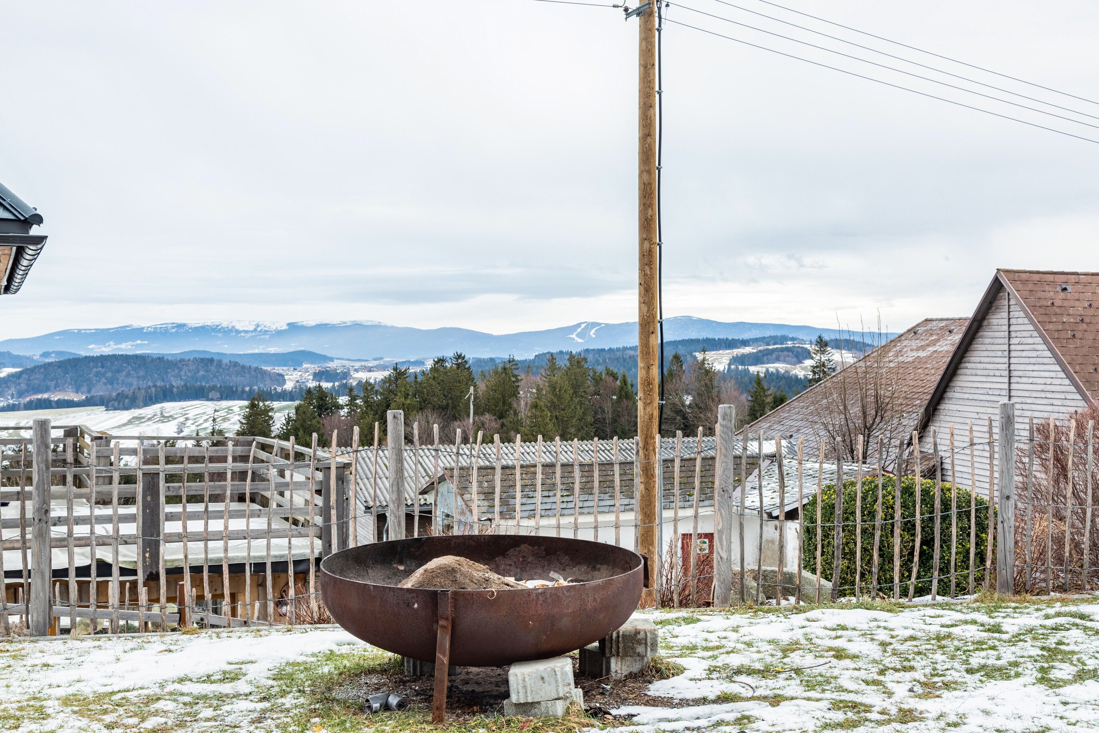 A metal fire pit in a snowy outdoor area with a wooden fence and a building in the background.