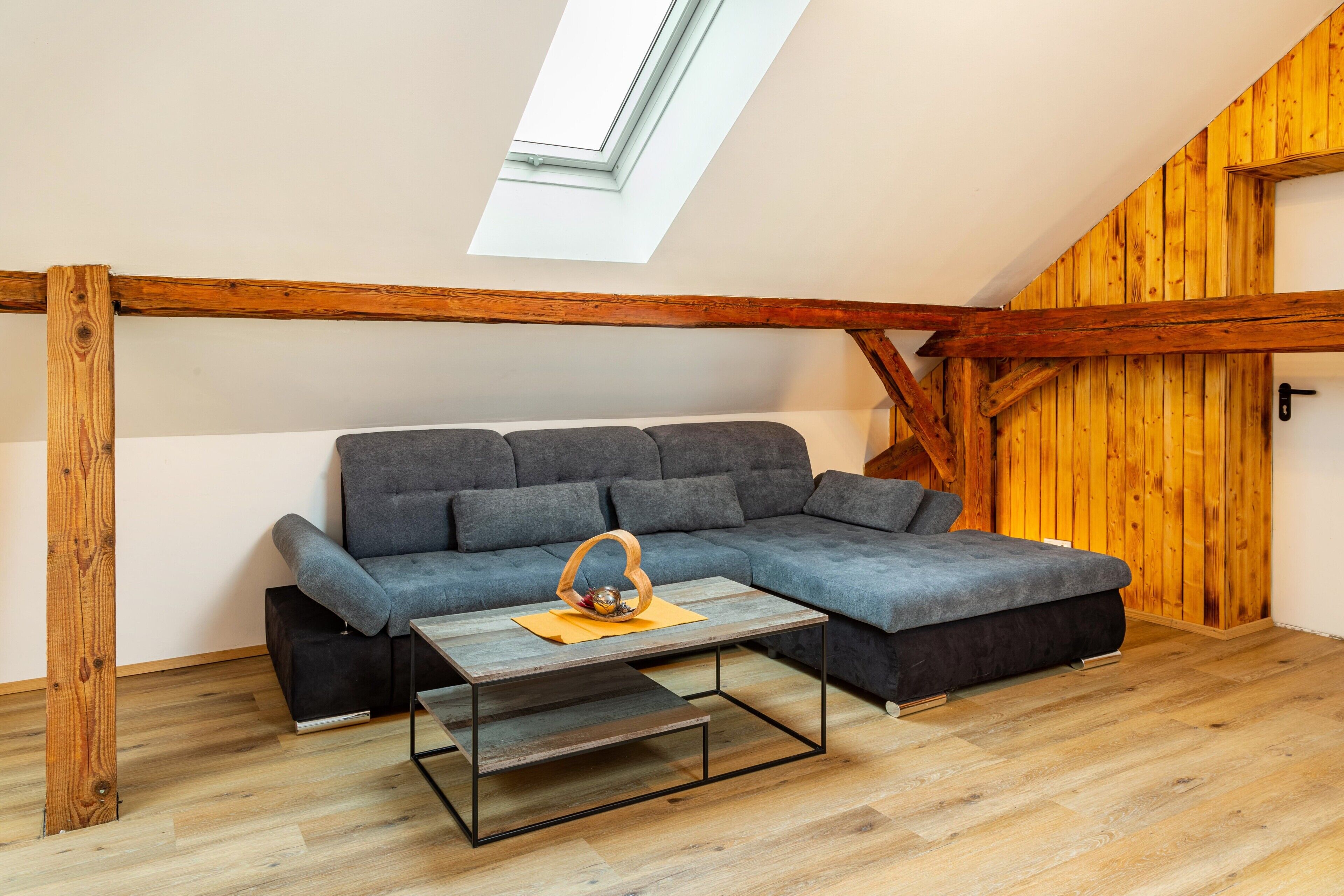Una habitación estilo loft con techo de viga de madera, sofá seccional gris, una mesita y una pared de madera.
