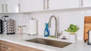 A kitchen with white cabinets, a stainless steel sink, a faucet, a coffee machine, a paper towel holder, a dish soap bottle, a sponge, and a potted plant. - Comfortable and Trendy Design 1BR in Lincoln Square (Chicago)