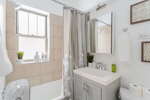 A modern bathroom with a bathtub, a toilet, a sink, and a window with plants. - Charming Guest Favorite 1BR in Lincoln Square (Chicago)