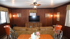 A room with a wooden wall paneling, a ceiling fan, a flat-screen TV mounted on a wooden cabinet, a brown leather sofa, an orange chair, and a dining table with a basket of flowers. - Entire Bear Den (cozy cabin) close to Glacier (Essex)