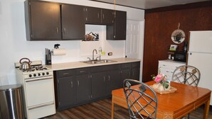 A compact kitchen with dark cabinets, a stainless steel sink, a white stove, and a dining table with metal chairs. - Entire Bear Den (cozy cabin) close to Glacier (Essex)