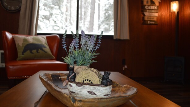 A wooden table with a rustic centerpiece, a leather sofa with a bear pillow, and a cabin-themed wall sign. - Entire Bear Den (cozy cabin) close to Glacier (Essex)