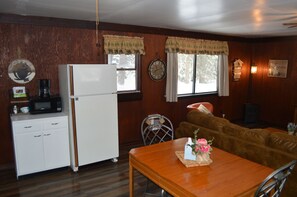 A compact kitchen with a refrigerator, microwave, and a coffee maker on the counter. There is a dining table with a vase of flowers and a brown leather couch in the living area.