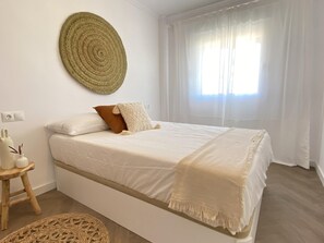 A bedroom with a bed, a round woven wall hanging, a bedside table with a vase, and a window with sheer curtains.