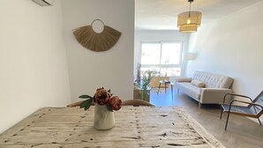 Une table à manger rustique, agrémentée d’un vase de fleurs, une décoration murale en bois et un canapé moderne dans un salon lumineux doté de grandes fenêtres.