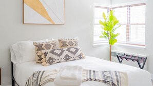 A neatly made bed with patterned pillows, a wall-mounted abstract painting, and a window with blinds and a green plant. - Modern Aesthetic 2BR in Evanston Near the Beach (Evanston)