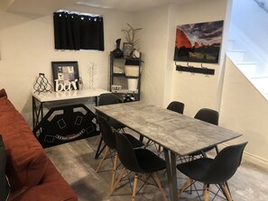 A dining area with a table and chairs, a black sofa, a framed picture, and a wall-mounted shelf with decorative items.
