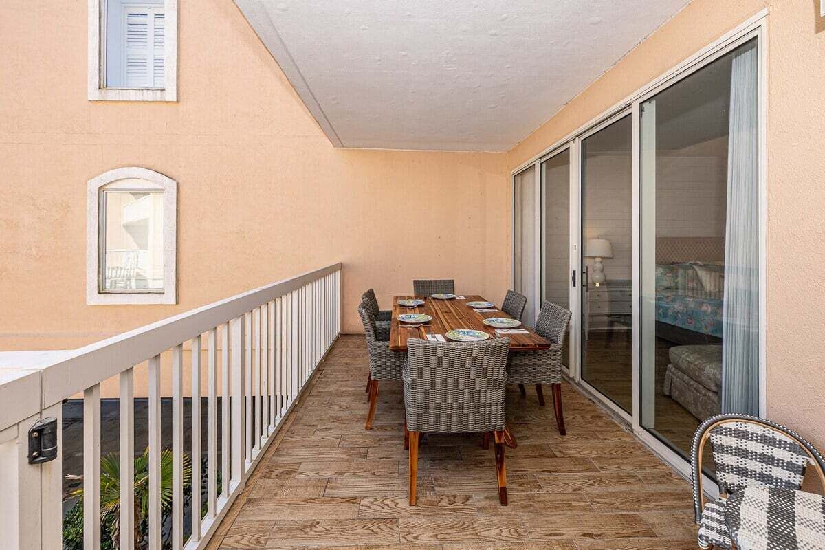 A balcony with a wooden table and wicker chairs, a white railing, and a sliding glass door leading to a room.