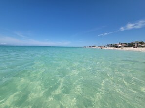 Ein Strand mit klarem türkisfarbenem Wasser und einigen Gebäuden im Hintergrund.