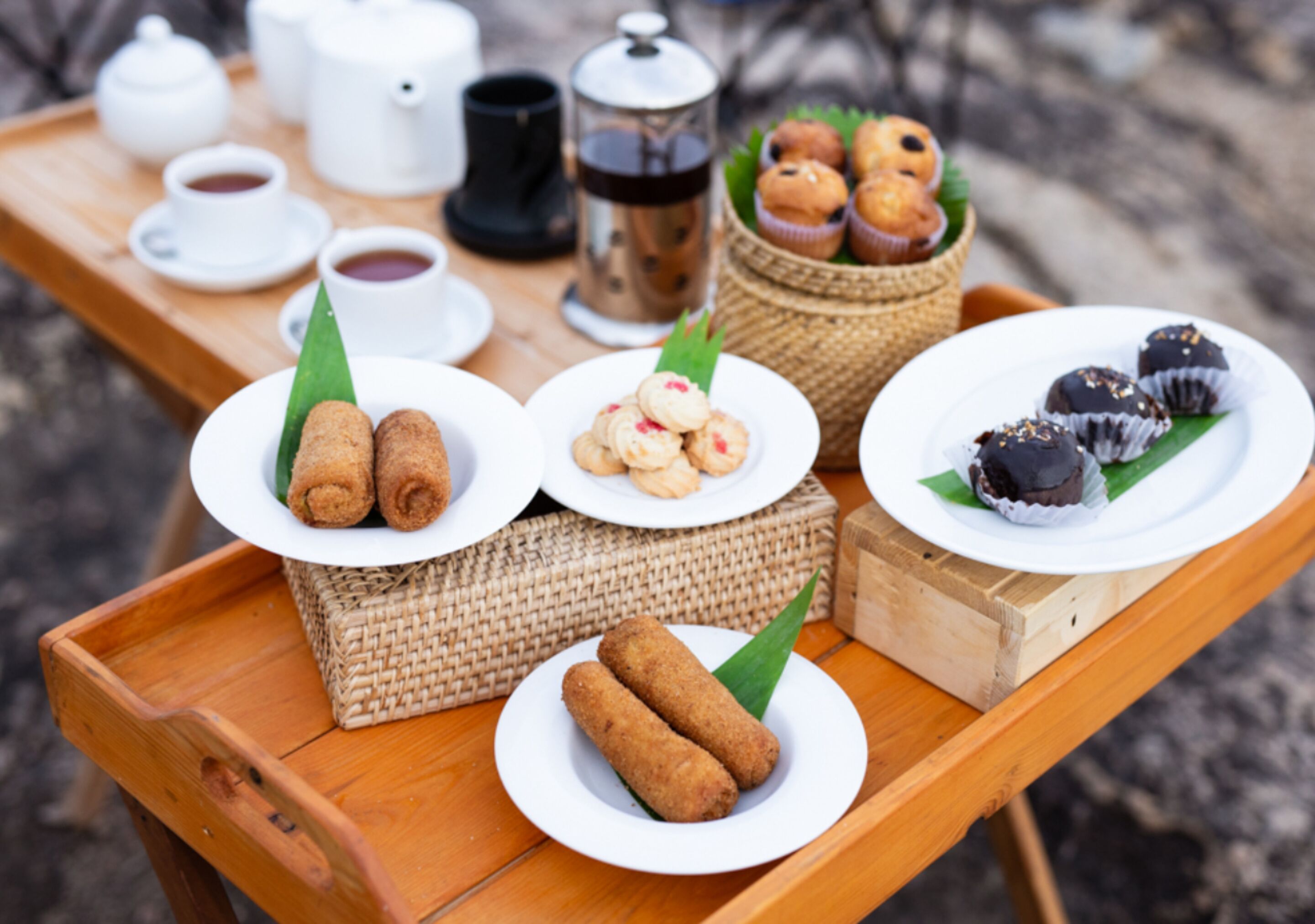 Una bandeja de madera con diversos alimentos, entre ellos croquetas, pasteles y dulces cubiertos de chocolate, sobre una mesa al aire libre.