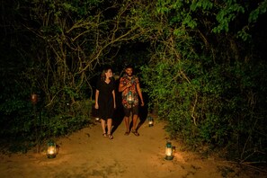 Two people walking on a dirt path surrounded by dense greenery, with lanterns placed along the path. - Tribe Yala - Luxury Camping (Thissamaharama)