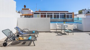Un área de piscina en la azotea con sillones de descanso y una mesa, rodeada de edificios.