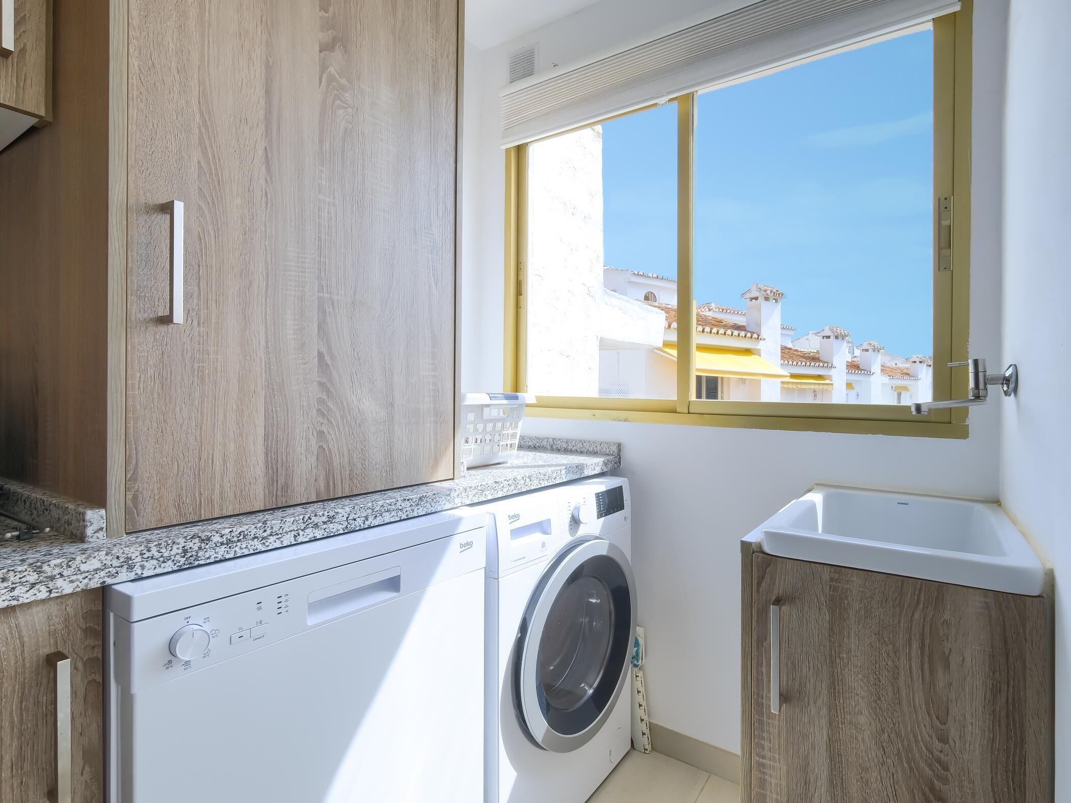 Una sala de lavandería con lavadora y secadora, armarios de madera, encimera de granito y ventana con vistas a edificios.