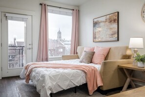 Una camera da letto con un letto, un divano, una finestra con tende, un quadro appeso e una lampada su un comodino.