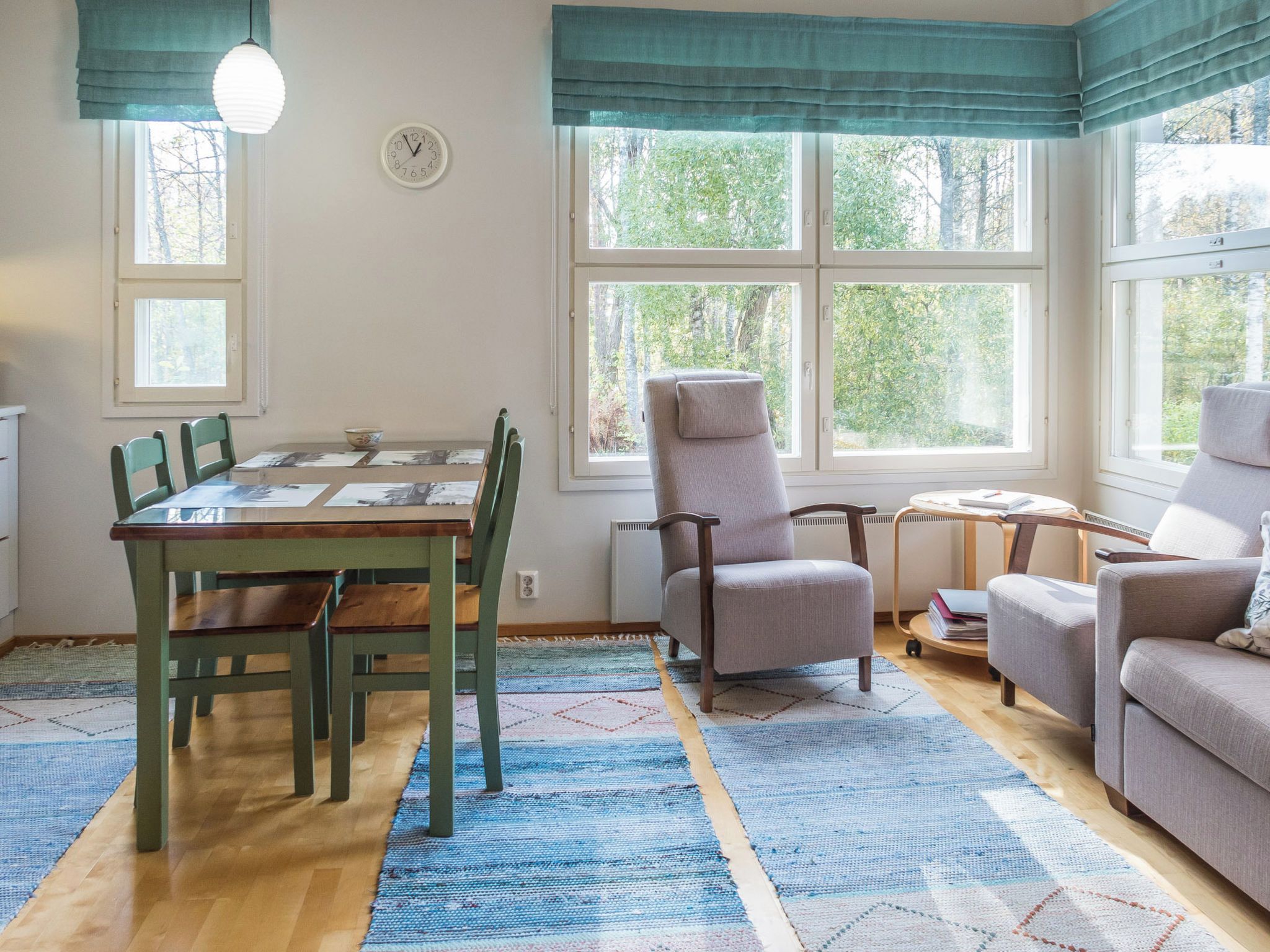 Huoneessa on ruokapöytä, tuoleja, sohva, nojatuoli, pieni pöytä, jossa on lamppu, ja seinällä oleva kello.
