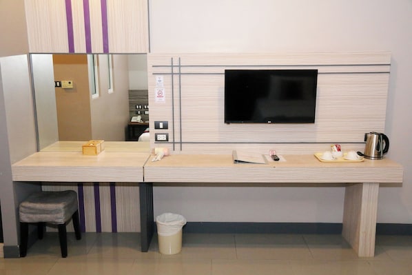 Habitación de hotel con escritorio, silla, televisor montado en la pared, bote de basura y cafetera.
