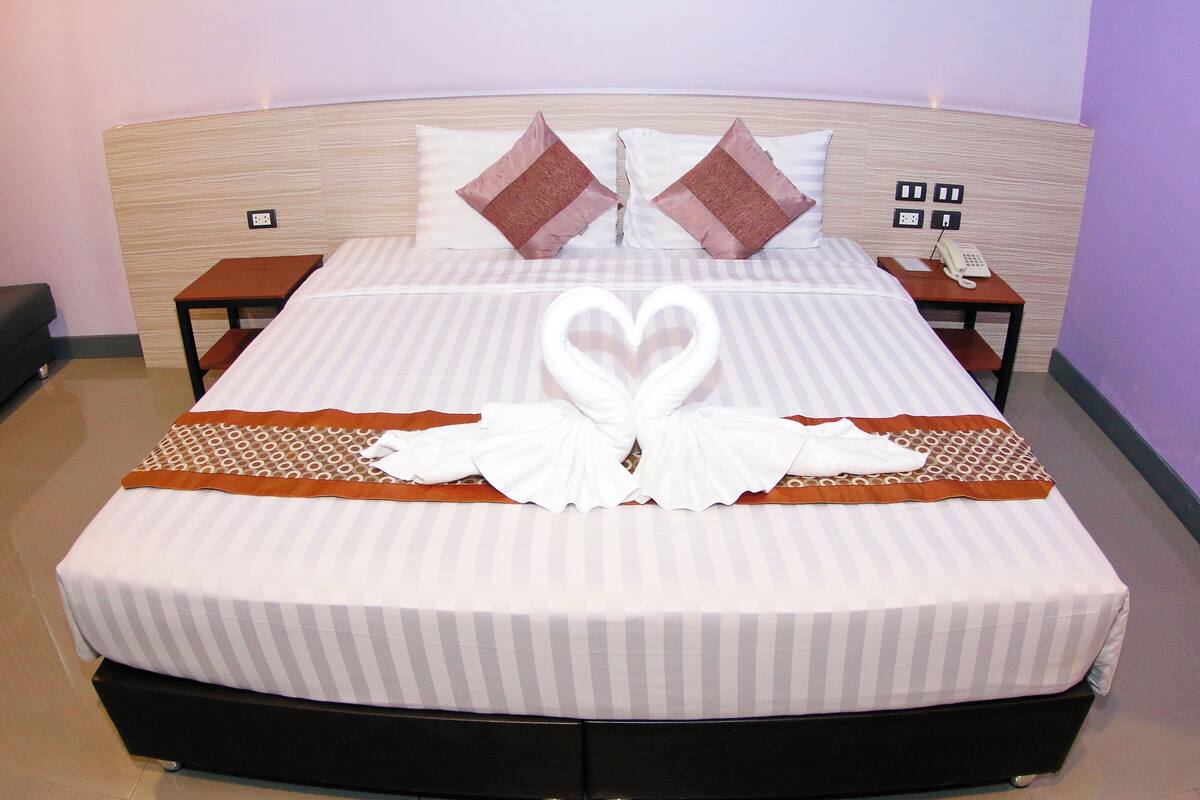 A neatly made bed with a heart-shaped towel arrangement, a wooden headboard, and bedside tables with a telephone and power outlets.