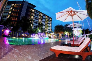 Zona de piscina nocturna con sillones de descanso y sombrillas, rodeada por un edificio con ventanas iluminadas.