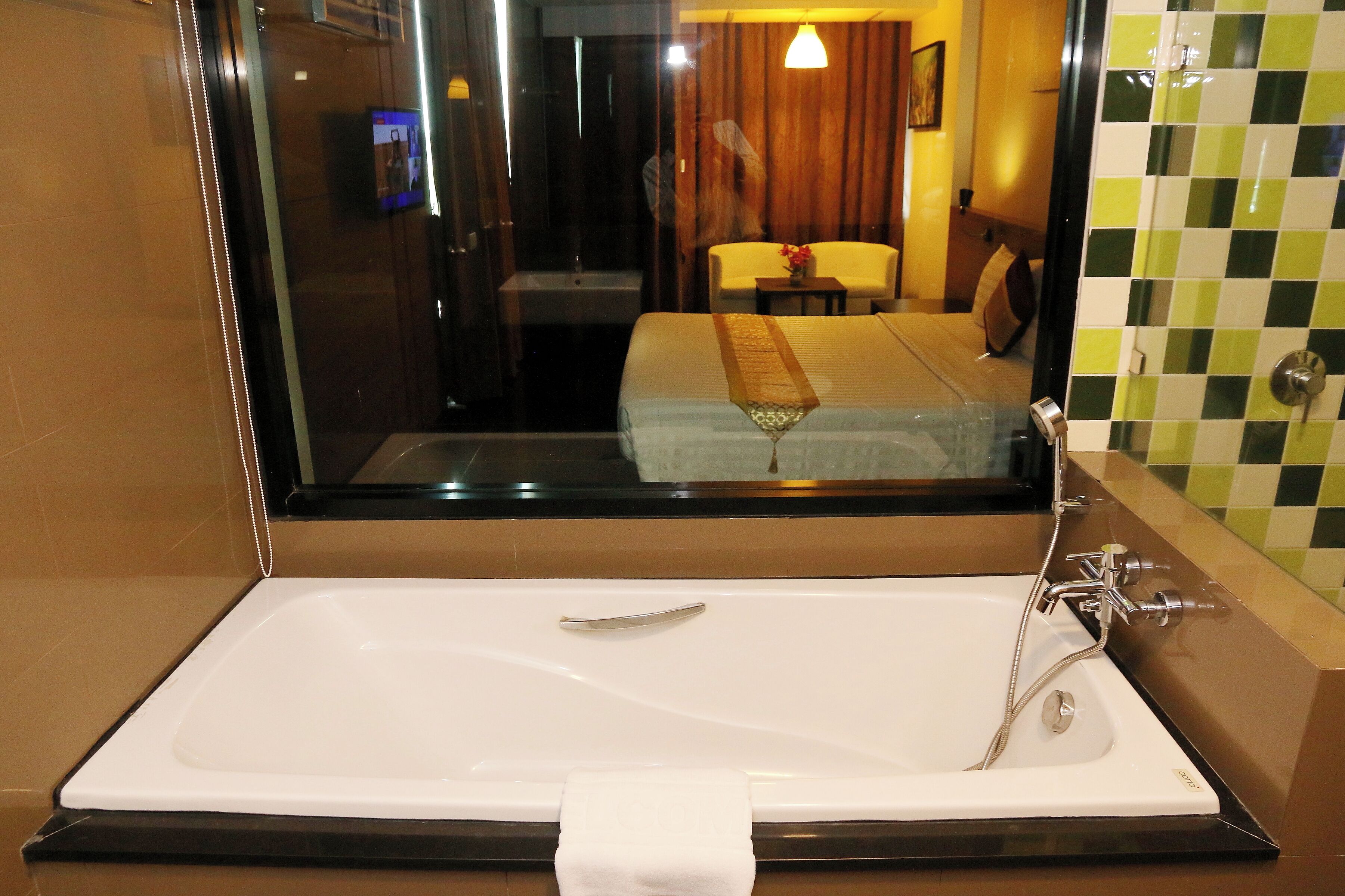 A hotel bathroom with a large bathtub, a mirror, and a view of a bedroom with a bed and a chair.