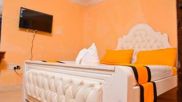 A bedroom with a white tufted bed, orange walls, and a flat-screen TV mounted on the wall.