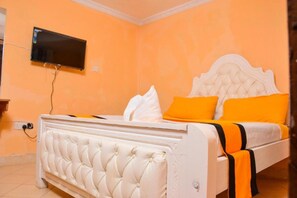 A bedroom with a white tufted bed, orange walls, and a flat-screen TV mounted on the wall.