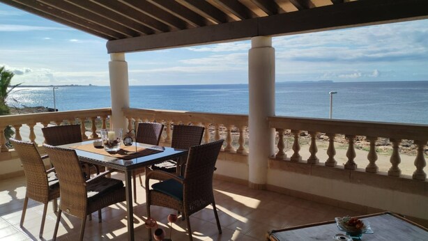 A covered outdoor dining area with a view of the sea, featuring a table with chairs and a small table with a vase.