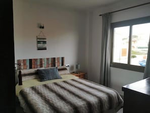 A bedroom with a bed, a bedside table, a lamp, a window with curtains, and a wall-mounted artwork.