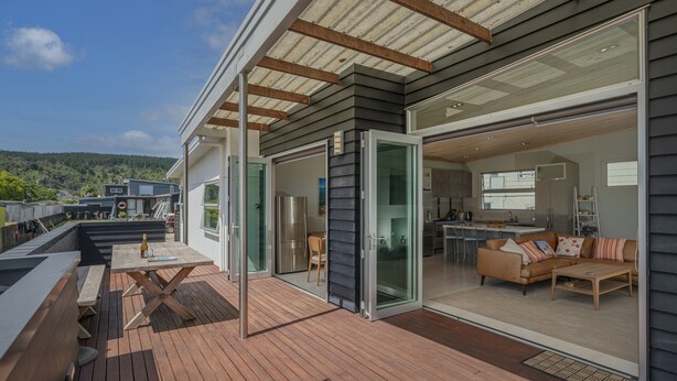 Een modern huis met een houten terras, schuiframen naar een woonkamer en uitzicht op het omliggende landschap.