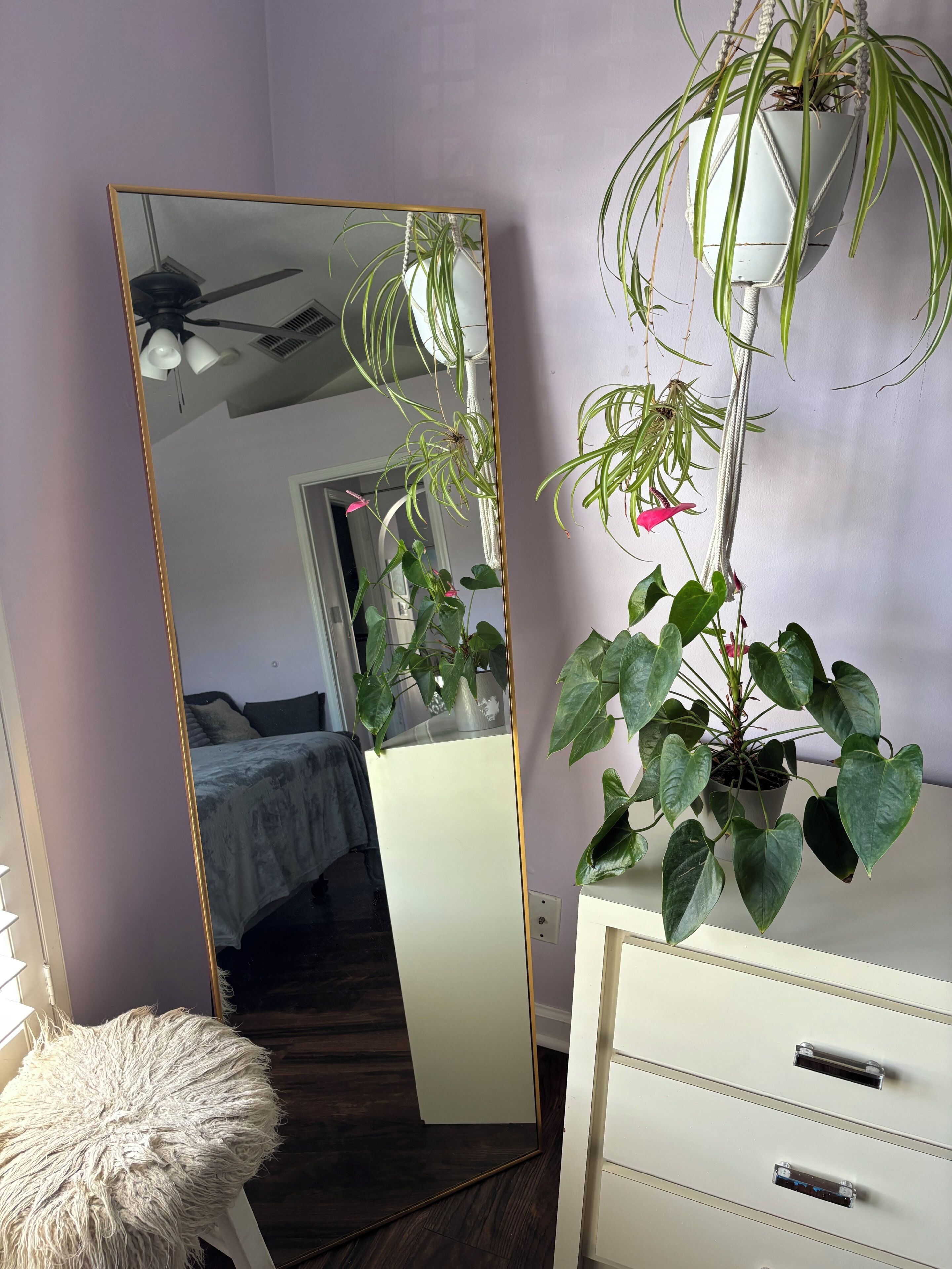 Een kamer met een grote spiegel die een plafondventilator weerspiegelt, een potplant met hartvormige bladeren, een pluizige witte kruk en een witte ladekast.