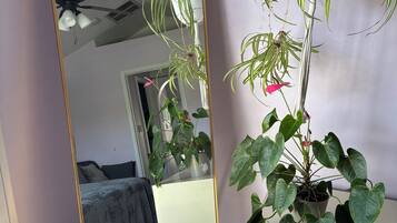 Een kamer met een grote spiegel die een plafondventilator weerspiegelt, een potplant met hartvormige bladeren, een pluizige witte kruk en een witte ladekast.