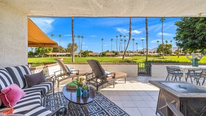 An outdoor patio with a sofa, chairs, and a table, overlooking a golf course with palm trees and houses in the background. - Modern Chic Condo at Rancho Las Palmas (Rancho Mirage)