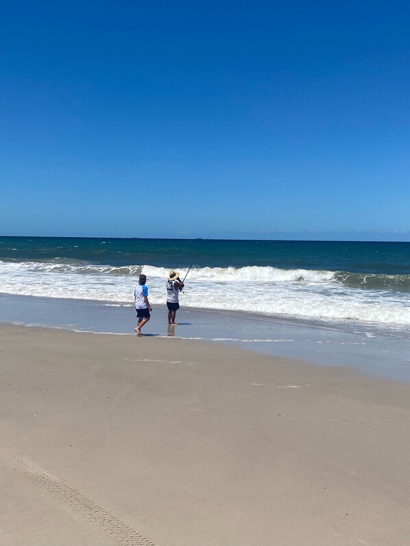 A beach with two people walking along the shore, one holding a fishing rod.