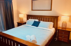 A neatly made bed with white linens and two rolled towels at the foot of the bed. There are two bedside tables with lamps, a wooden headboard, and a wall-mounted picture frame.