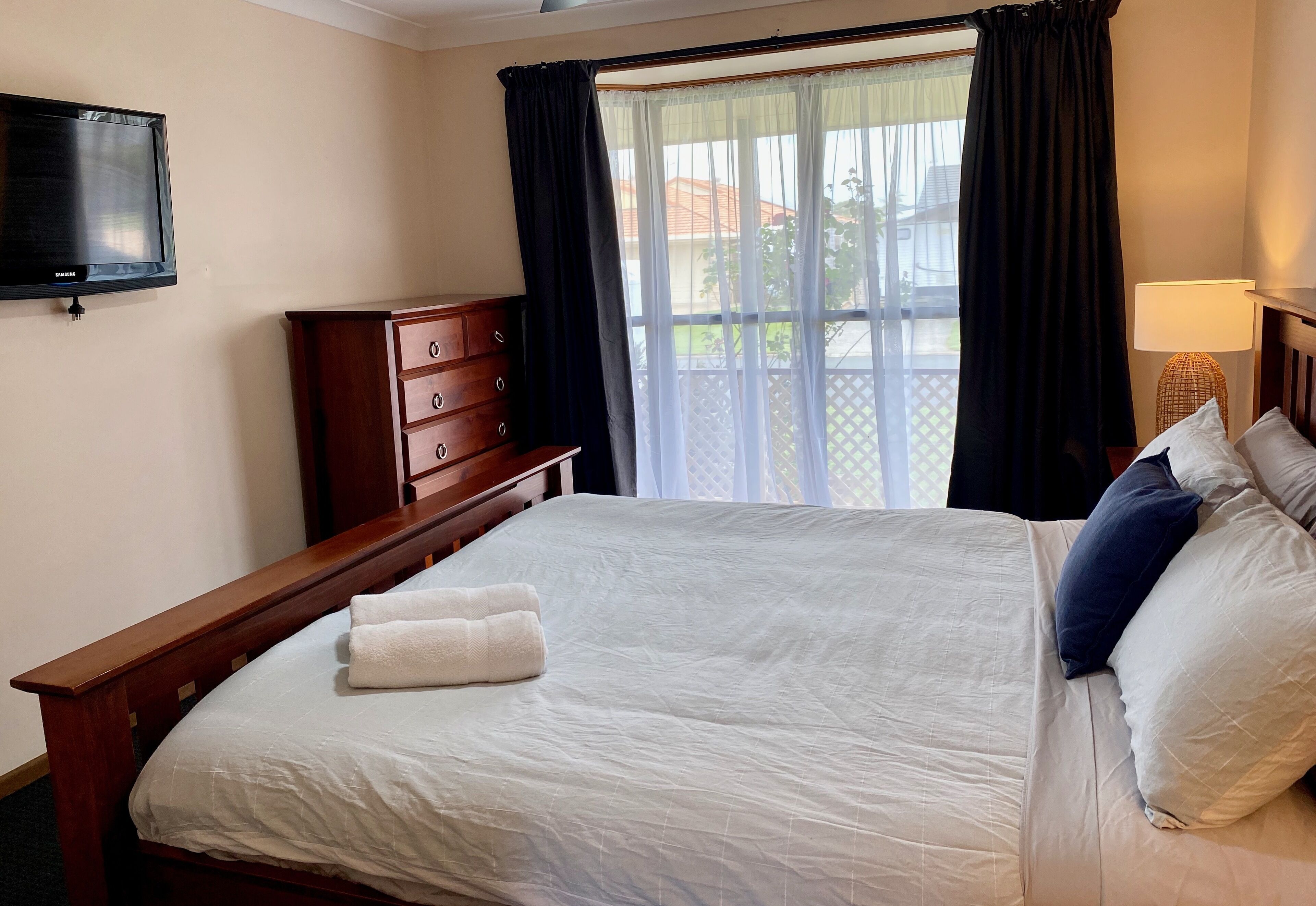 A neatly made bed with white linens and a folded towel, a wooden dresser, a flat-screen TV mounted on the wall, and a window with black curtains.