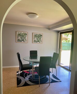 A dining area with a glass table and four chairs, two framed tropical leaf artworks on the wall, and a view of a green fence through a sliding glass door.