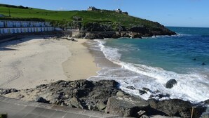 Una platja costanera amb un litoral rocos, un turó verd amb una construcció, i un cel blau clar.