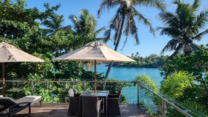 A wooden deck with outdoor furniture and umbrellas overlooking a body of water, surrounded by lush greenery and palm trees.