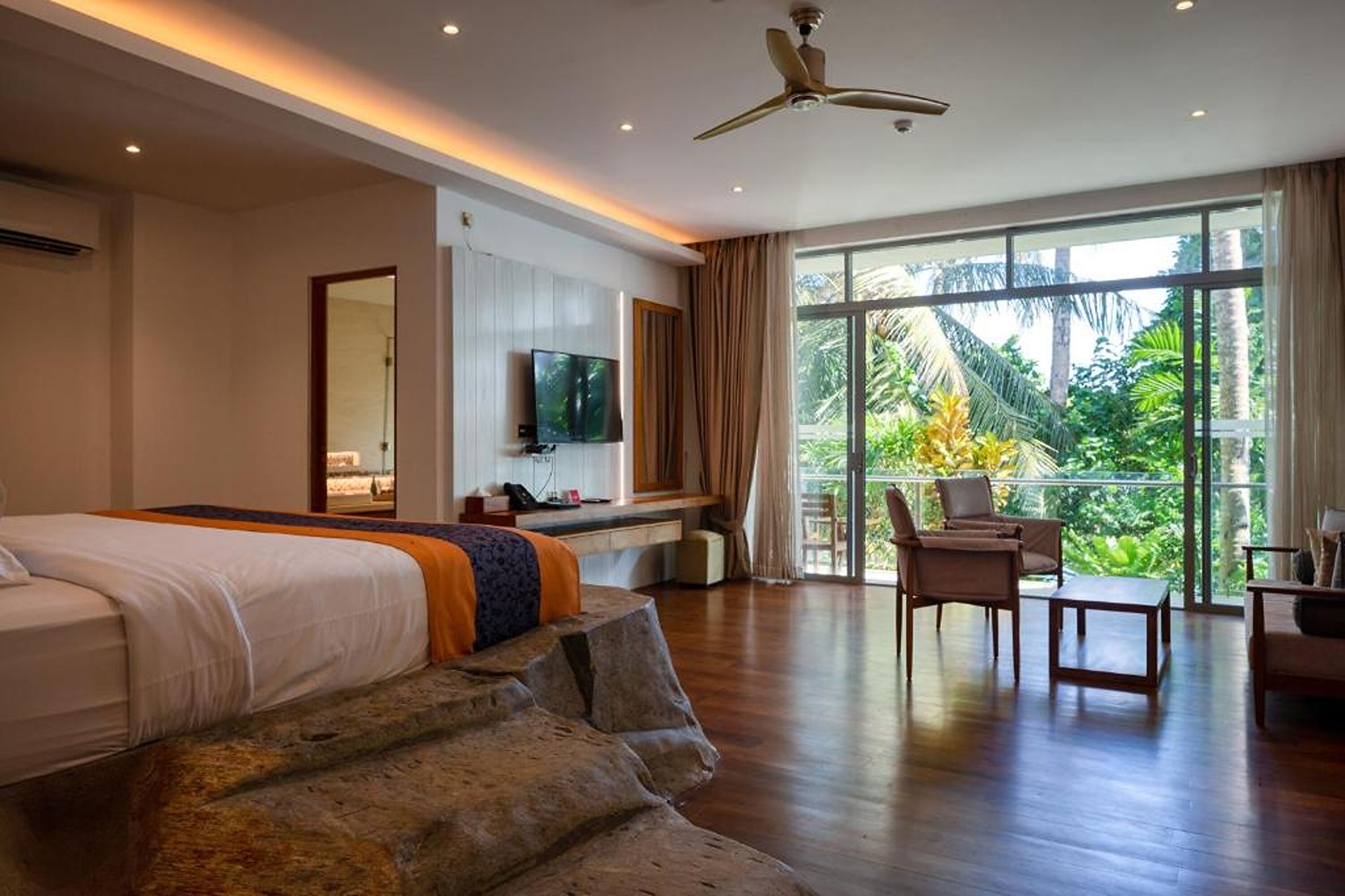 A modern hotel room with a large bed, a flat-screen TV, a balcony with a view of lush greenery, and a ceiling fan.