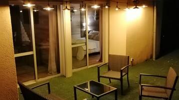 An outdoor patio area with a glass enclosure, furnished with chairs and a table, and illuminated by hanging lights.