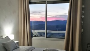A hotel room with a large bed, two pillows, a nightstand, a lamp, and a view of the mountains through a window with curtains. - Lapausedegout (Lachapelle-Auzac)
