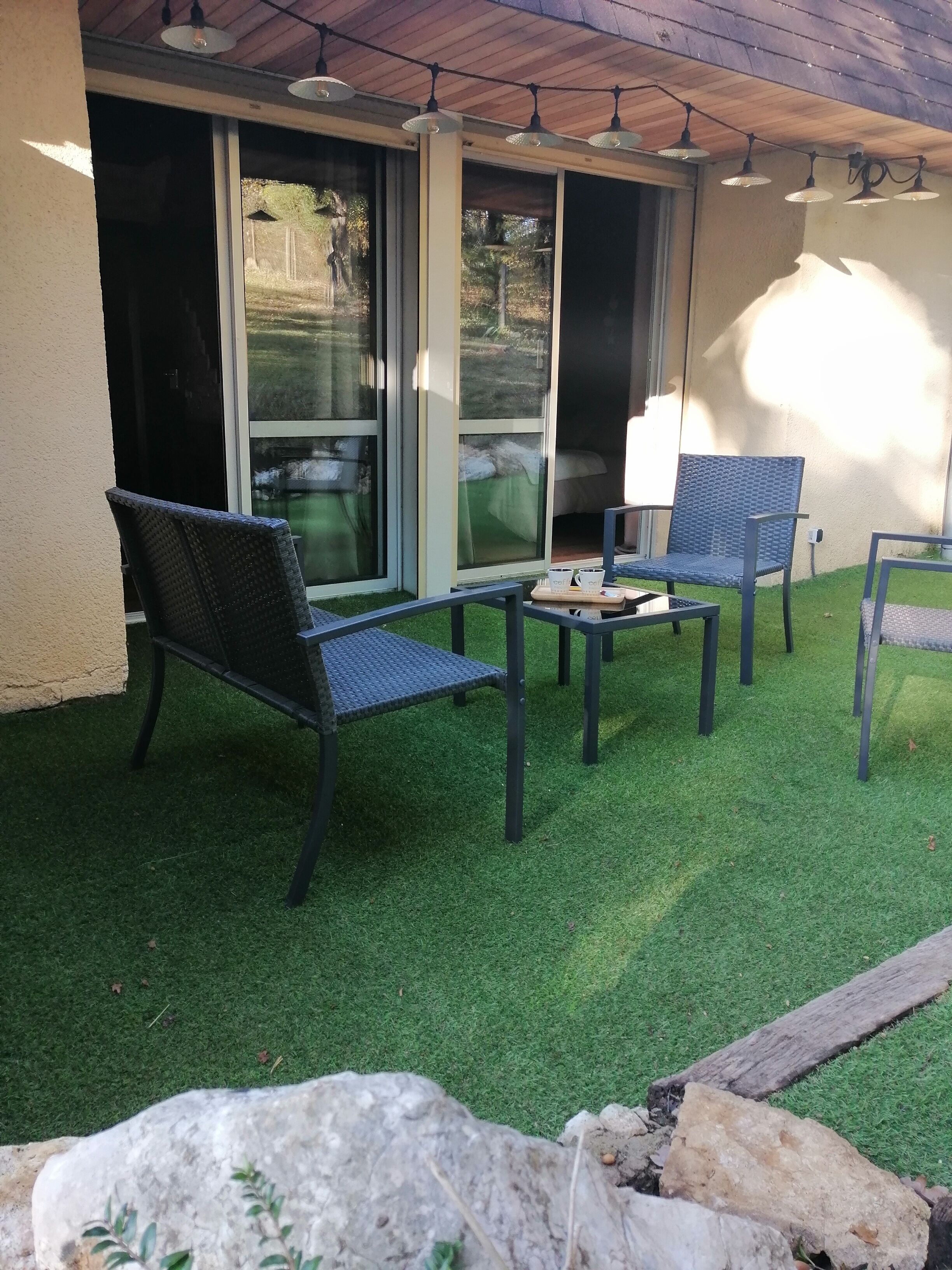 Un patio al aire libre con césped artificial, dos sillas de mimbre, una mesita y una guirnalda de luces.