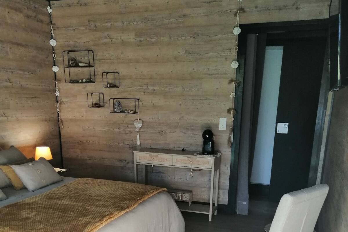 Une chambre avec un mur en bois, un lit recouvert d’une couverture beige, une table de chevet avec une lampe et une chaise.