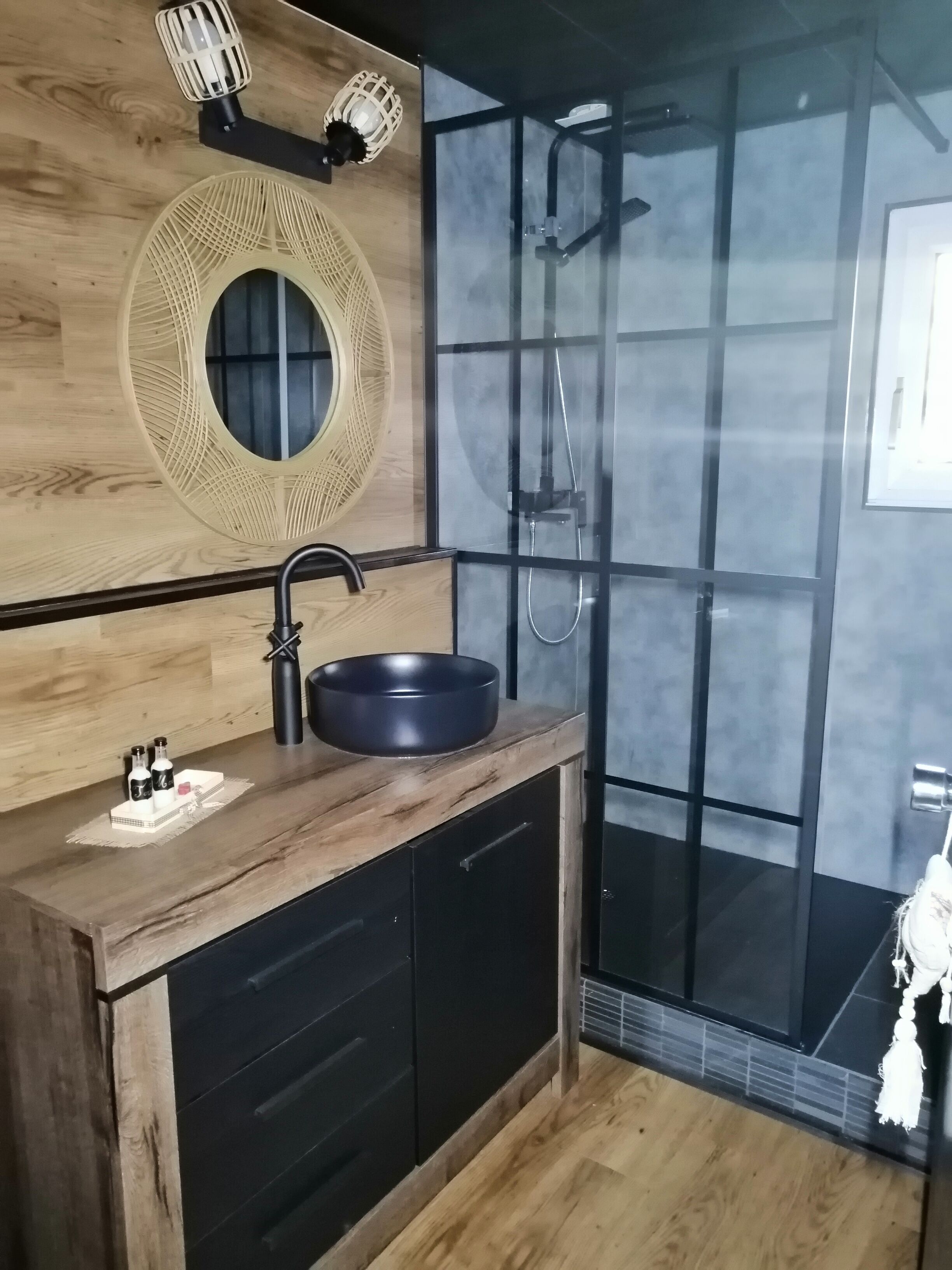 A modern bathroom with a wooden vanity, a round mirror, a black sink, and a glass shower enclosure.